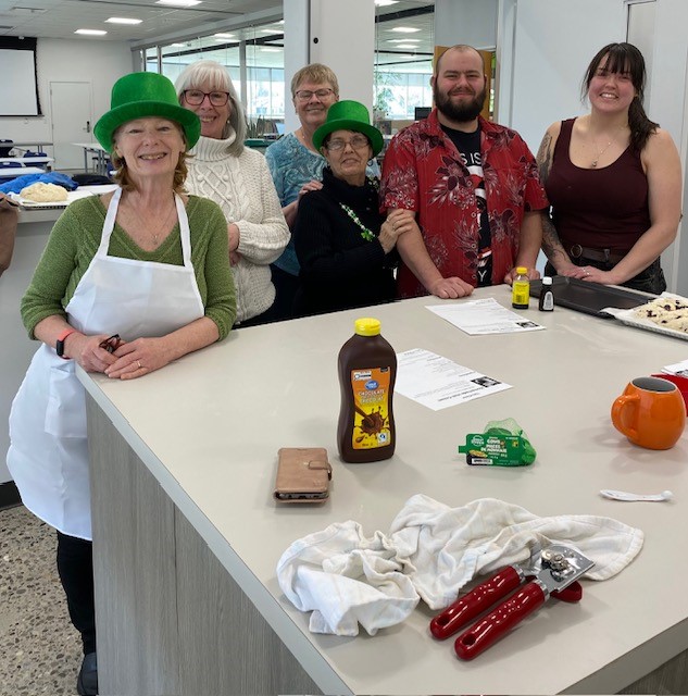 group of adults at a table with cooking supplies, two are wearing green top hats