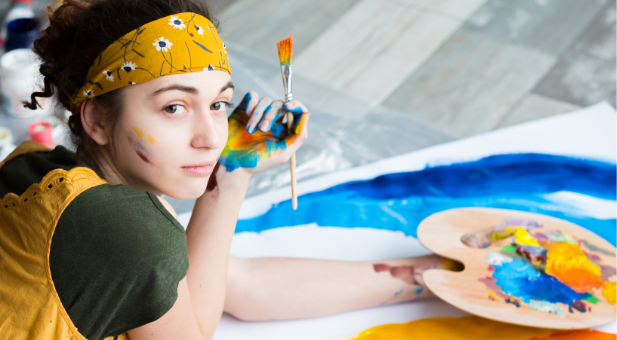 teen girl wearing a yellow bandana and holding a paint brush looks at the camera